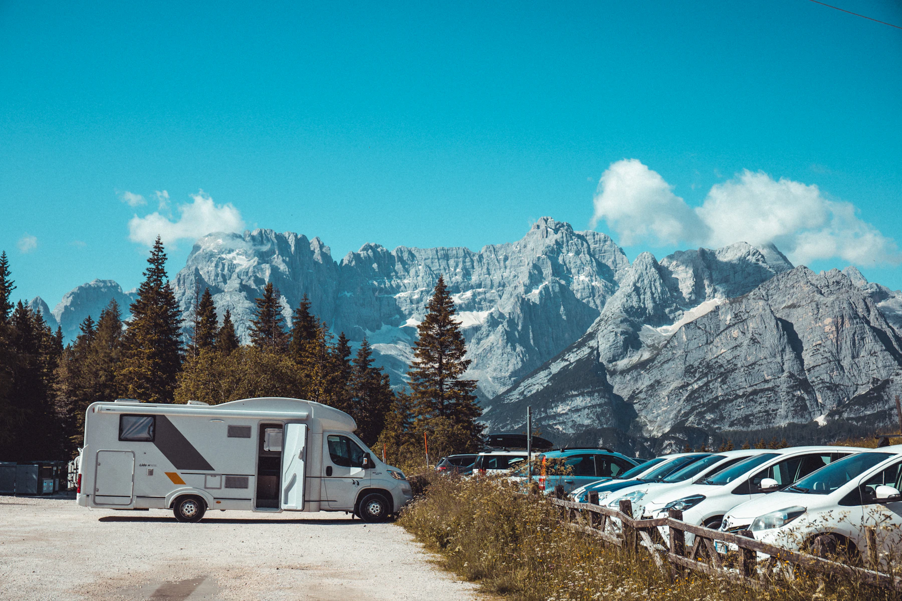 Motorhomes and cars parked in a gravel stopover below jagged mountains