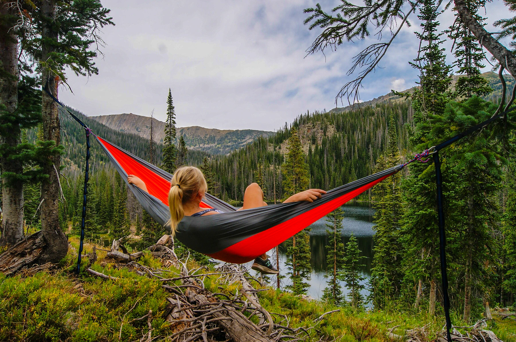 A person resting in a hammock strung between pines above a still mountain lake